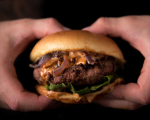 A pair of hands holding a fresh juicy burger with lettuce onions and cheese on black background - Menu Alphaville